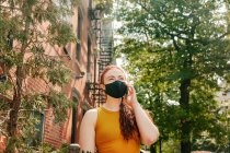 Jeune femme au téléphone portant un masque de visage dans la rue Brooklyn par les arbres — Photo de stock