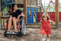 Улыбающаяся мать в инвалидном кресле смотрит на свою маленькую дочь, играющую в — стоковое фото