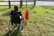 Маленькая девочка играет в зеленом поле со своей матерью в инвалидной коляске — стоковое фото