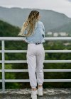 Back side of a young woman with blonde braided hair wearing a denim jacket and white jean resting on a dam — Stock Photo