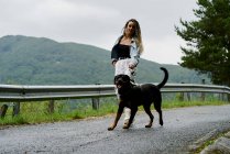 Giovane donna con capelli biondi intrecciati che indossa una giacca di jeans e jeans bianco che porta a spasso il suo cane in una giornata piovosa — Foto stock