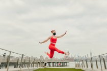 Young female athlete jumping mid air wearing face mask by waterfront — Stock Photo