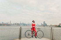 Молодая велосипедистка в маске для лица с велосипедом на набережной — стоковое фото