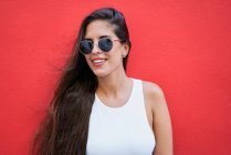 Jovem mulher com cabelos longos vestindo óculos de sol elegantes e de pé perto da parede do edifício vermelho na cidade — Fotografia de Stock