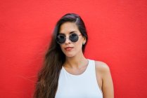 Giovane donna con i capelli lunghi indossa occhiali da sole alla moda e in piedi vicino al muro rosso edificio in città — Foto stock