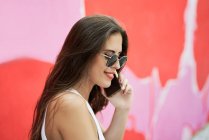Молода жінка в сонцезахисних окулярах і розмовляє на мобільному телефоні, сидячи на сходах зі стіною пофарбованою в рожевий і червоний колір на задньому плані. Концепція технології — стокове фото
