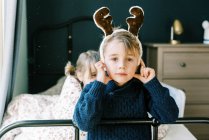 Petit garçon dans un pull en tricot bleu avec un chapeau de renne dans la chambre sur le lit — Photo de stock