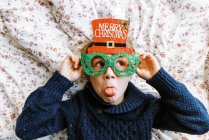 Маленький мальчик в вязаном свитере в рождественских очках лежит на кровати — стоковое фото