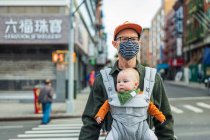Father wearing face mask carrying daughter in baby carrier while crossing street in city during pandemic — Stock Photo