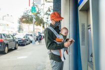 Отец в маске для лица носит дочь в детской коляске во время использования кредитной карты в банкомате во время пандемии — стоковое фото