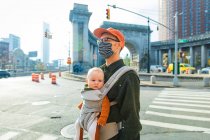 Vater trägt Tochter in Tragetasche, während er während der Coronavirus-Pandemie auf der Straße in der Stadt läuft — Stockfoto