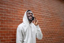 Plan latéral de l'homme africain avec geste heureux et capuche en utilisant un smartphone en ville — Photo de stock