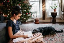 Jovem mulher lendo com seu cão pela árvore de Natal — Fotografia de Stock
