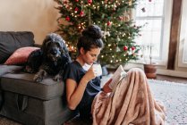 Jeune femme lecture et boire du thé avec chien bu arbre de Noël — Photo de stock