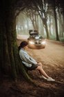 Jovem mulher encostada à árvore musgosa na floresta, carro no fundo — Fotografia de Stock