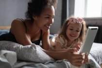 Задоволена етнічна жінка і дівчина лежать на ліжку і грають в гру на цифровому планшеті разом у вихідні вдома — стокове фото