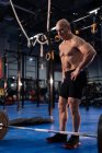 Selbstbewusster älterer Sportler mit den Händen auf der Taille, der neben der Langhantel steht und sich ausruht, während er vor dem Gewichtheben im Fitnessstudio posiert — Stockfoto