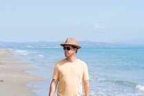 Man with hat and sunglasses standing on seashore, while looking away — Stock Photo