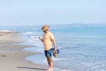 Чоловік стоїть на березі моря, тримаючи взуття і спостерігаючи оболонку — стокове фото
