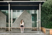 Young girl sitting alone on a wooden seat at an empty bus stop — Stock Photo