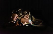 Irmãos lendo um livro debaixo de um cobertor por lanterna à noite. — Fotografia de Stock