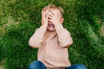 Glücklicher kleiner Junge lächelt und bedeckt sein Gesicht, während er im Gras liegt — Stockfoto