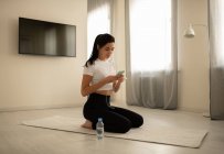 Vista laterale di bruna bella donna seduta su un materassino con auricolari che navigano nel cellulare durante l'allenamento a casa — Foto stock