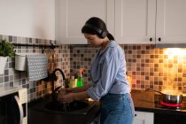 Vue latérale de la jeune femme au foyer en chemise bleue et jeans écoutant de la musique avec écouteurs sans fil et lave-vaisselle dans la cuisine à la maison avec de l'eau bouillante dans la casserole en arrière-plan — Photo de stock