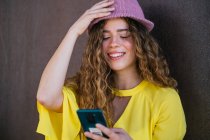 Entzückte Frau passt stylischen Hut an, während sie Smartphone benutzt — Stockfoto