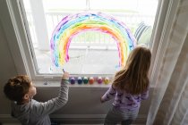 Сверху вниз вид братьев и сестер, рисующих радугу на окне гостиной — стоковое фото