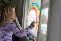 Вид сбоку улыбающейся девушки, рисующей радугу на окне — стоковое фото