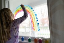 Visão traseira da menina loira pintando arco-íris na janela — Fotografia de Stock