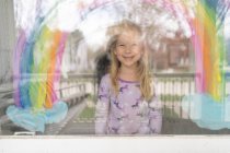 Lächelndes blondes junges Mädchen unter Regenbogen auf Fenster gemalt — Stockfoto