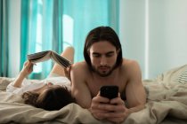 Жінка читає книгу на ліжку біля чоловіка, використовуючи смартфон, відпочиваючи вранці вдома — стокове фото