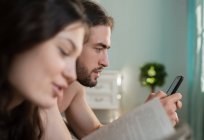 Bärtiger Rüde nutzt Smartphone, während er morgens zu Hause neben lesenden Weibchen ruht — Stockfoto