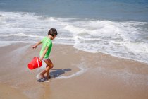 A little girl with a bucket plays in the tide at edge of sandy beach — Stock Photo