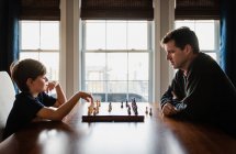 Father and son sitting at a table indoors playing a game of chess. — Stock Photo
