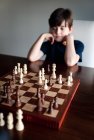 Jovem menino pensativo sentado atrás de um tabuleiro de xadrez olhando para as peças. — Fotografia de Stock