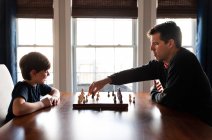 Père et fils assis à une table à l'intérieur jouant à un jeu d'échecs. — Photo de stock
