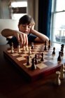 Nachdenklicher Junge sitzt hinter Schachbrett und bewegt eine der Figuren. — Stockfoto
