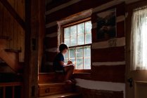 Niño sentado en los escalones de una cabaña de madera mirando por la ventana - foto de stock