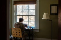 Мужчина сидит за столом перед окном в спальне и работает за компьютером. — стоковое фото