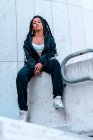 Молода чорна жінка сидить з косами та татуюваннями, дивлячись сидячи — стокове фото
