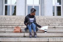Estudiante africana universitaria usando mascarilla protectora estudiando sentada en escaleras afuera en el campus. Nuevo normal en la universidad. - foto de stock