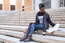 Eine afrikanische Studentin mit Schutzmaske sitzt draußen auf dem Campus auf einer Treppe. Neue Normalität im College. — Stockfoto