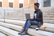 Estudiante africana universitaria usando mascarilla protectora estudiando en su portátil sentada en escaleras afuera en el campus. Nuevo normal en la universidad. - foto de stock