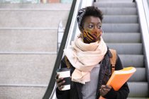 Estudiante africana universitaria con máscara protectora subiendo escaleras mecánicas a la estación de metro. Nueva normalidad en el transporte público. - foto de stock