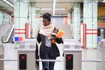 Африканская студентка университета в защитной маске проходит через турникеты с транспортной картой на станции метро. Новая норма в общественном транспорте. — стоковое фото