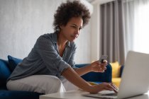 Mujer negra usando portátil y tarjeta de crédito para hacer compras en línea en el sofá en casa - foto de stock