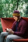 Joven macho usando su smartphone descansando en una hamaca en el bosque - foto de stock